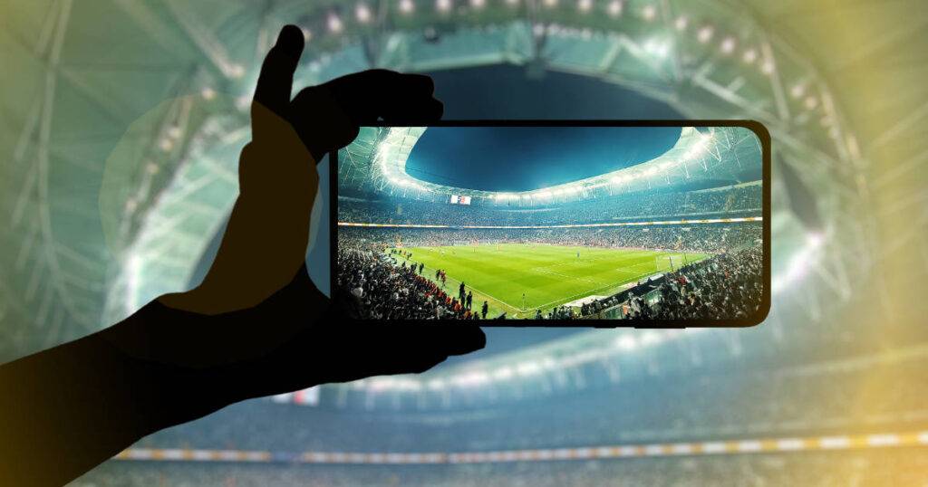 Fan hand with mobile phone horizontal focused on sports field showcasing global fan engagement in sports.